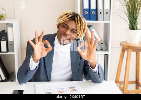 Great job. Happy black man. Successful achievement. Stylish positive guy sitting office desk showing ok gesture feeling satisfied in light room interi Stock Photo
