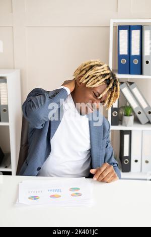 Office work. Sick black man. Headache suffer. Exhausted grimacing from pain guy holding hand on neck sitting desk light room interior. Stock Photo