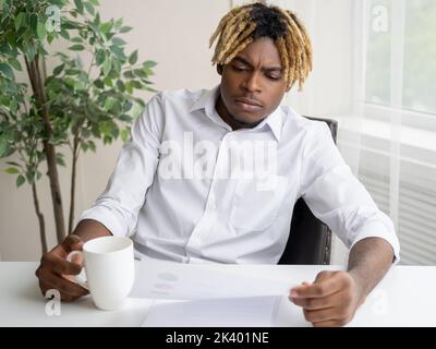 Office routine. Gloomy black man. Checking documents. Handsome serious guy sitting work desk with cup of coffee looking paper information in light roo Stock Photo