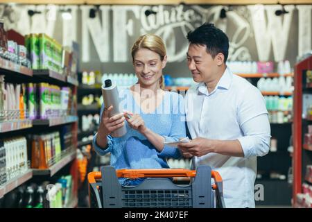 A young interracical couple, a woman and an Asian man walk between the rows with a trolley in a supermarket, the woman holds a bottle of shampoo in her hands, reads the composition on the label. Stock Photo
