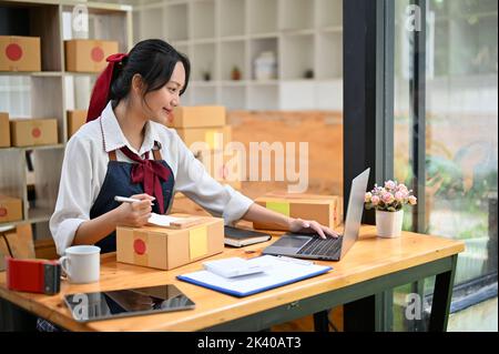 Pretty and charming young Asian female small online business owner checking her orders on laptop while preparing shipping boxes. Stock Photo