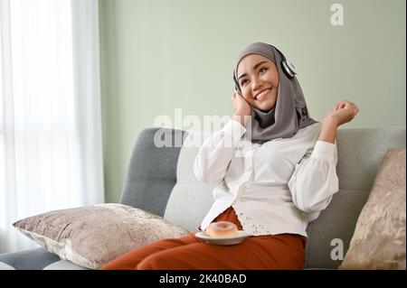 Joyful and happy young Asian Muslim woman wearing hijab listening to music though her smartphone while relaxing in her living room. Stock Photo