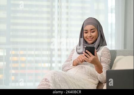 Attractive and beautiful millennial Asian Muslim pregnant woman resting in her home living room, using her mobile phone on the sofa. Motherhood concep Stock Photo