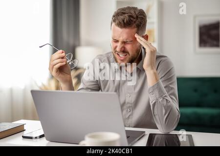 Young bearded coder with headache, handsome man has burnout at workplace with laptop, tired male working from home Stock Photo