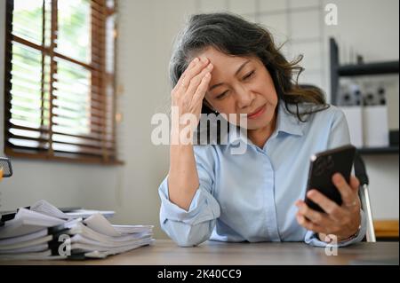 Stressed and sad Asian aged businesswoman or senior female manager at her desk, staring at her smartphone screen, receiving bad news, having problems Stock Photo