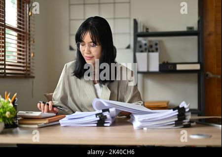 Professional and charming millennial Asian businesswoman or female accountant wearing eyeglasses, working at her desk, calculating financial and accou Stock Photo