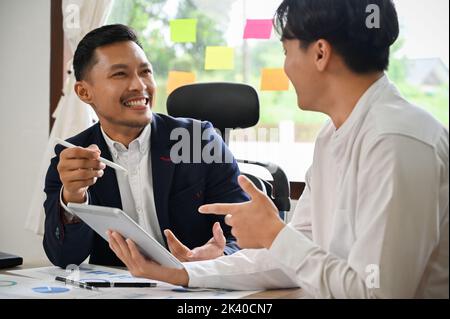 Two Asian male business coworkers or businessmen enjoy chatting, sharing business ideas while working together on their project. Stock Photo