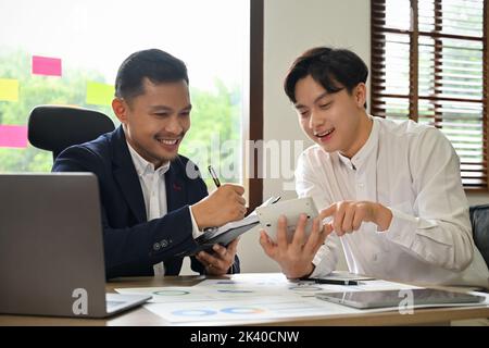 Handsome and cheerful young Asian businessman or male employee having a nice conversation with his colleague during work together. Stock Photo