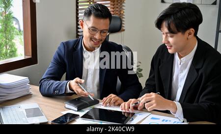 Smart and professional adult Asian businessman giving advices and explaining something to his male staff while working together in the office. Stock Photo