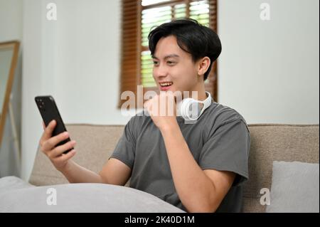 Handsome and happy young Asian man using his smartphone while relaxing on a comfy sofa in his living room. reading an online blog, social media, chatt Stock Photo