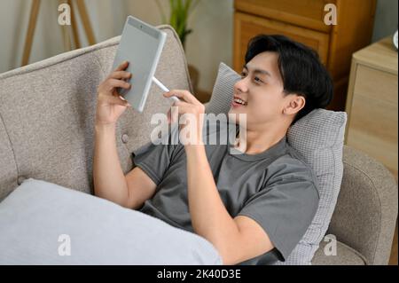 Handsome young Asian man relaxes lying on the sofa and enjoys reading online fiction or cartoons on his tablet. Stock Photo