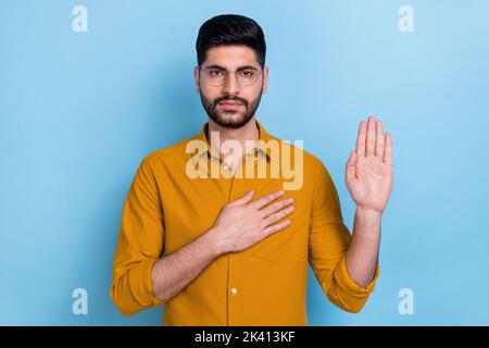 Portrait of calm focused young person in yellow shirt glasses arm touch heart raise palm swear isolated on blue color background Stock Photo