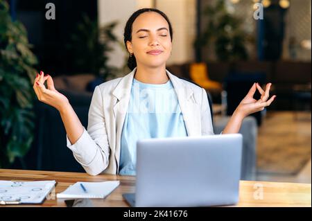 Positive calm relaxing beautiful young mixed race woman, office worker, company manager, sitting at workplace in the office, resting from work, meditating with closed eyes, smiling Stock Photo