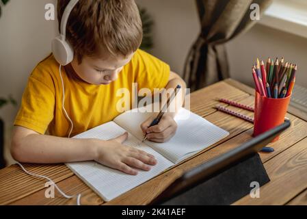 The child is a boy in a yellow T-shirt with headphones doing homework on a tablet. The concept of remote school education at home. Stock Photo