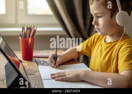 The child is a boy in a yellow T-shirt with headphones doing homework on a tablet. The concept of remote school education at home. Stock Photo