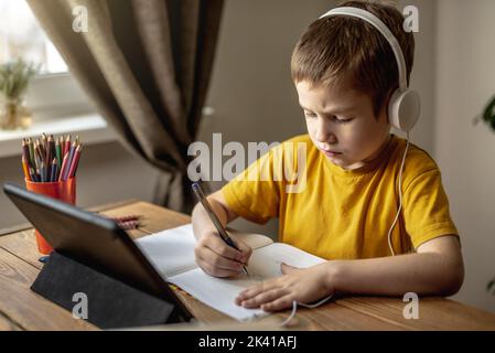 The child is a boy in a yellow T-shirt with headphones doing homework on a tablet. The concept of remote school education at home. Stock Photo