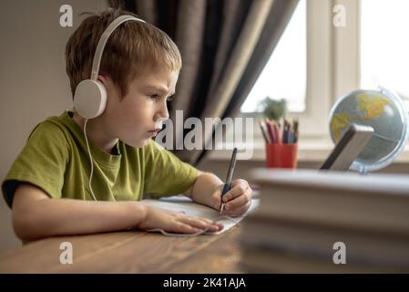 The child is a boy in a yellow T-shirt with headphones doing homework on a tablet. The concept of remote school education at home. Stock Photo