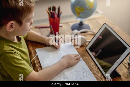 The child is a boy in a yellow T-shirt with headphones doing homework on a tablet. The concept of remote school education at home. Stock Photo