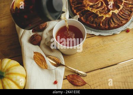 Cozy autumn. Pouring herbal tea in stylish cup on background of wooden table with freshly baked warm apple pie with cinnamon, autumn leaves, pumpkins Stock Photo