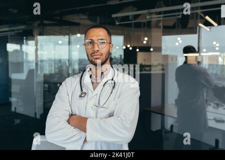 Portrait of serious and successful doctor, man in medical gown looking thoughtfully at camera, working inside modern office building. Stock Photo