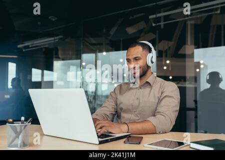 Young office worker working inside office building, using laptop to watch video and headphones, businessman on break. Stock Photo