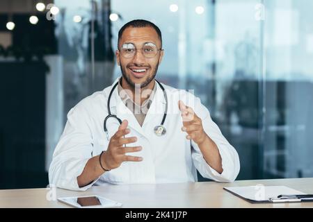 Webcam view, cheerful and successful family doctor consults patients remotely online, video call of man in medical coat smiling and looking at camera, working inside clinic office. Stock Photo
