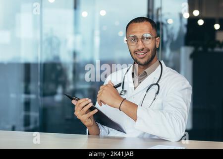 Webcam view hispanic doctor looking at camera and consulting patients man in medical gown working remotely inside modern clinic office. Stock Photo