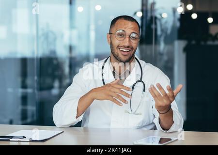 Webcam view, cheerful and successful family doctor consults patients remotely online, video call of man in medical coat smiling and looking at camera, working inside clinic office. Stock Photo