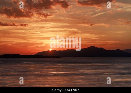 Offshore view of sun setting behind mountains as sky glows orange Stock Photo
