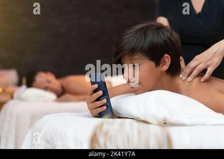 Child is using the phone and having a massage in a spa with his mother Stock Photo