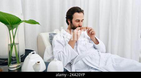 Portrait of handsome man in white bathrobe drinking healing tea while relaxing in wellness center. Men spa program Stock Photo