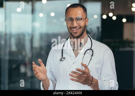 Close-up photo of hispanic doctor smiling and looking at web camera talking to colleagues and patient on video call, man in medical coat and stethoscope working inside clinic office. Stock Photo
