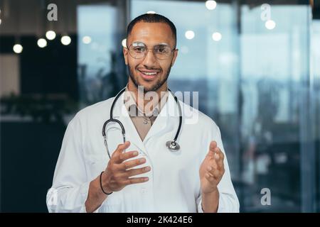 Close-up photo of hispanic doctor smiling and looking at web camera talking to colleagues and patient on video call, man in medical coat and stethoscope working inside clinic office. Stock Photo