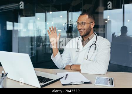 Happy hispanic doctor smiling and talking on video call man in white coat and stethoscope using laptop for remote online consultation smiling and waving hello. Stock Photo