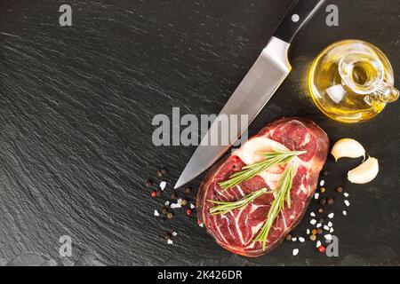 Fresh raw meat beef steak with bone with spices, rosemary, knife and olive oil on the black slate surface. cooking ingredients. Top view. Flat lay. Stock Photo