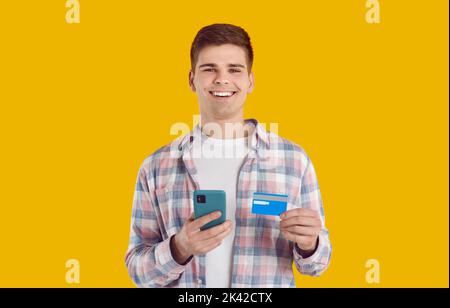 Young Caucasian man smiling holds phone and credit card for shopping stands in yellow studio Stock Photo