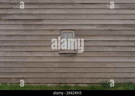 Rustic wood cladding texture with small centred wood framed window, horizontal unpainted old wood planks background, grass and plants at the base Stock Photo