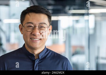 Close up photo portrait of successful young Asian investor, man in glasses smiling and looking at camera in shirt, businessman working inside modern office building, satisfied accountant financier. Stock Photo