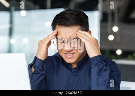 Close-up photo of sick Asian businessman having severe headache, male boss in glasses working inside modern office building, holding hands on head massaging, overtired at work. Stock Photo