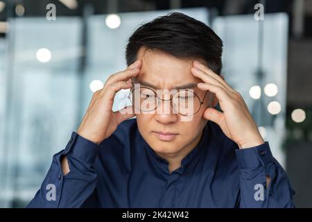 Closeup thinking businessman, Asian man in glasses and shirt holding hands on head making important decision, male manager working inside modern office building, thoughtful and serious boss. Stock Photo