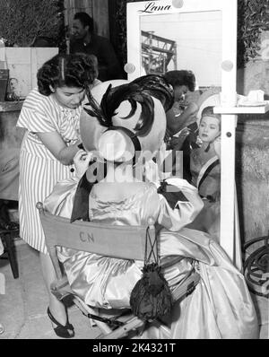 LANA TURNER puts on a bonnet in front of her personal mirror assisted by Hairdresser HELEN YOUNG on set location candid in Northern California during filming of GREEN DOLPHIN STREET 1947 director VICTOR SAVILLE novel Elizabeth Goudge screenplay Samson Raphaelson women's costumes Walter Plunkett Metro Goldwyn Mayer Stock Photo