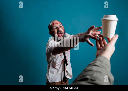 Spooky horror monster wanting cup of coffee, chasing after hand with carton cup with drink. Aggressive scary zombie eating brain and looking dangerous with bloody scars or wounds. Stock Photo