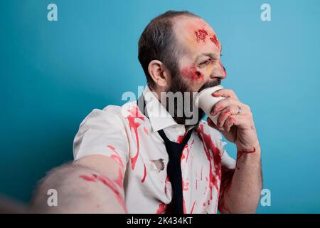 Dangerous halloween zombie biting on coffee cup and acting aggressive, looking creepy and taking photo in studio. Undead evil monster corpse with bloody scars eating carton, doomsday. Stock Photo