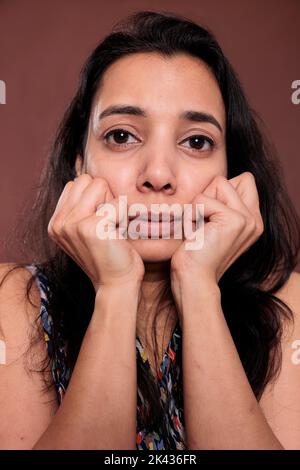 Indian woman holding clenched fists on cheeks closeup portrait, cute pose. Calm lady touching face with hands close view, person with neutral facial expression looking at camera Stock Photo