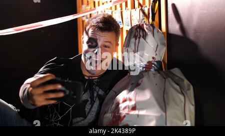 Halloween party, night, twilight, in the rays of light, a man with a terrible makeup does selfie with a corpse wrapped in oilcloth, the corpse is smeared with blood. High quality photo Stock Photo