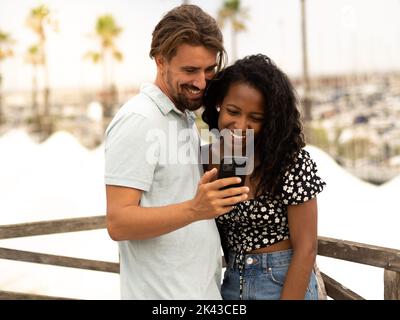Diverse couple of happy heterosexual lovers looking at smartphone in a city on vacation Stock Photo