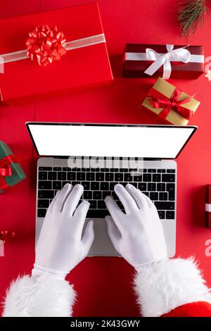 Image of hands of santa claus using laptop with blank screen and gifts on red background Stock Photo
