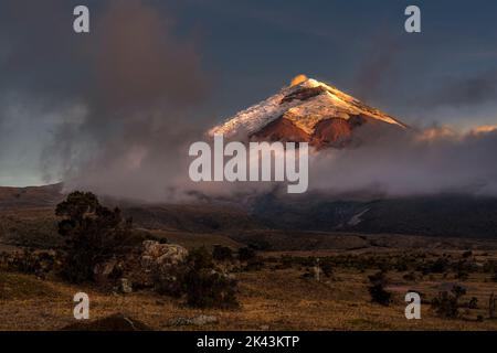 Cotopaxi volcano at dusk. The full moon is rising on the left lighting the volcano meanwhile the orange light of the setting sun is illuminating  the Stock Photo
