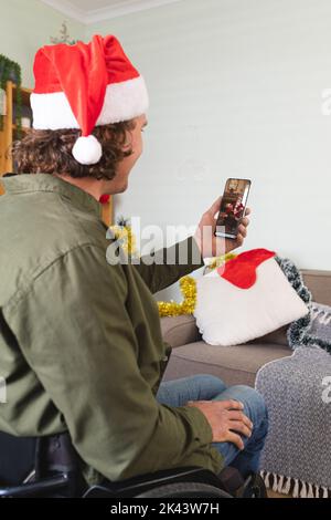 Caucasian man in wheelchair and santa hat making smartphone christmas video call with santa claus. christmas, festivity and communication technology. Stock Photo