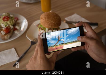 Hands of caucasian man at restaurant watching female athlete doing long jump on smartphone. sports, competition, entertainment and technology concept Stock Photo